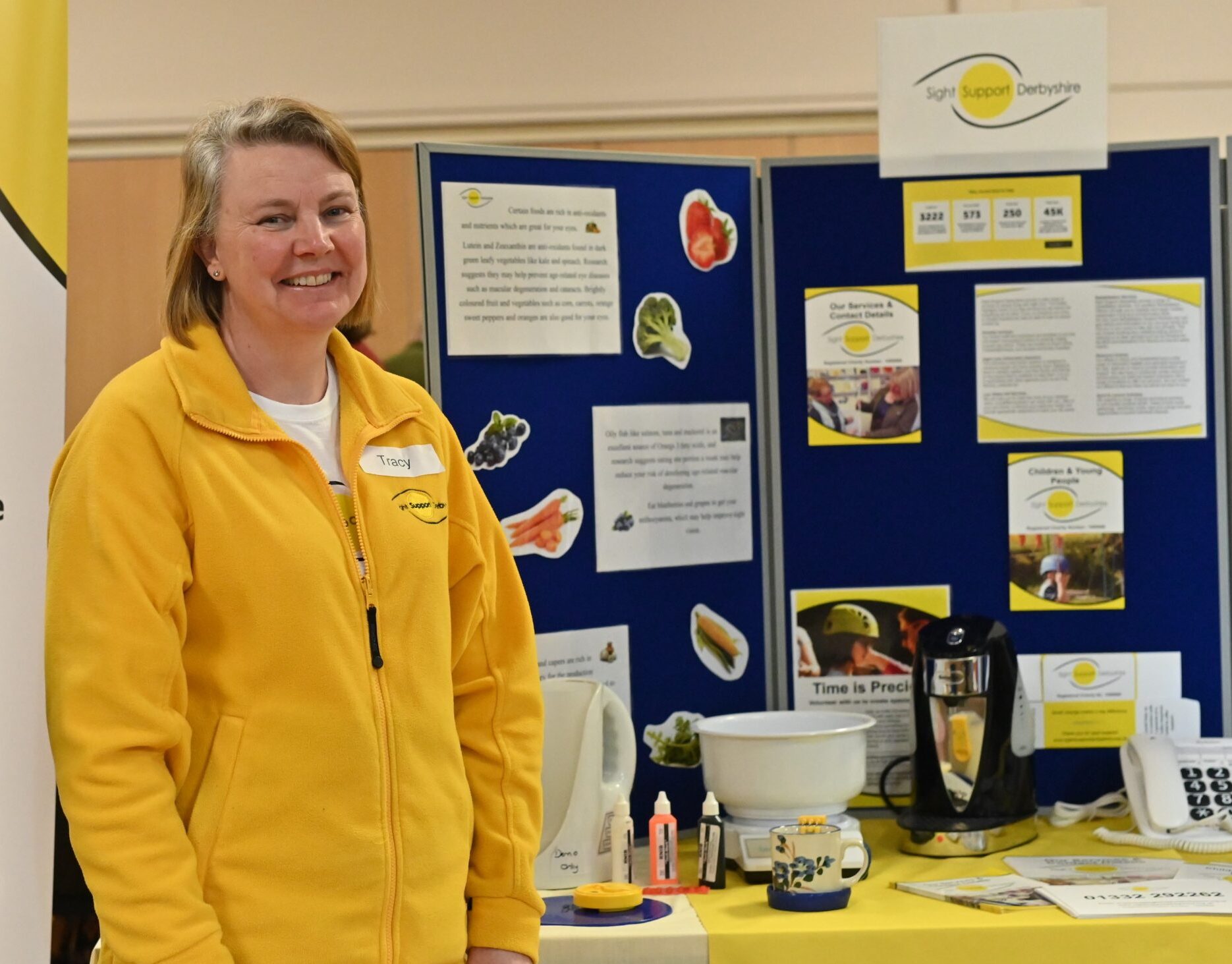 A lady wearing a yellow Sight Support Derbyshire fleece is standing next to a display board and table with equipment. She is smiling at the camera