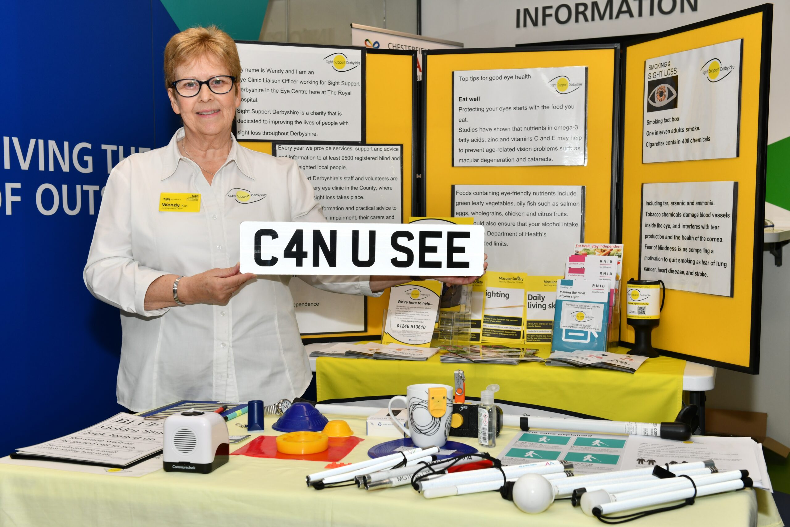 Wendy is wearing a white shirt and is standing in front of a display of leaflets, equipment (talking clocks, canes etc). She is holding a care registration plate which reads C4N U SEE