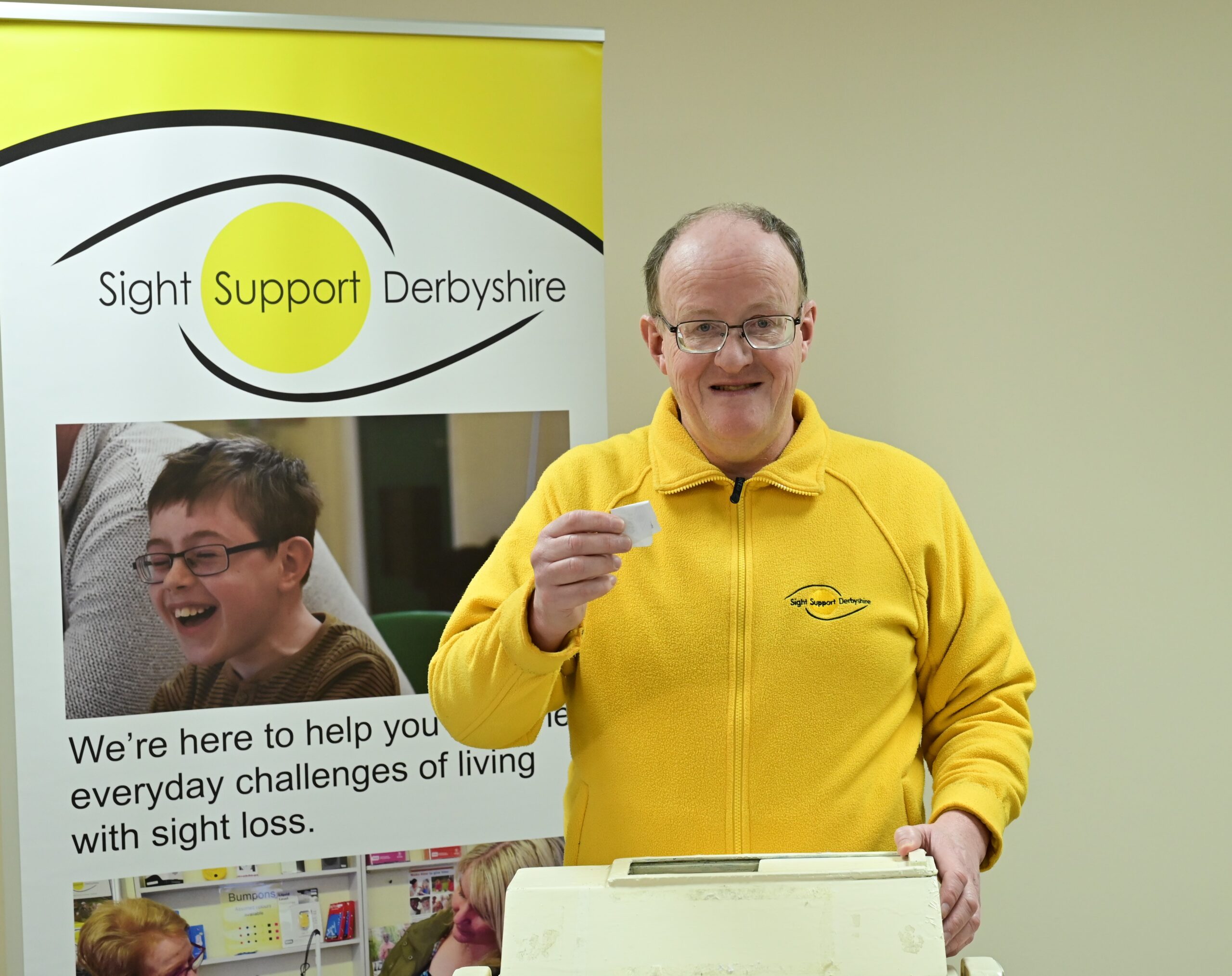 A gentleman wearing a bright yellow fleece with the Sight Support Derbyshire logo is smiling and holding up a raffle ticket which he has just pulled out from a 'tombola' drum. To the left of the picture is a Sight Support Derbyshire banner