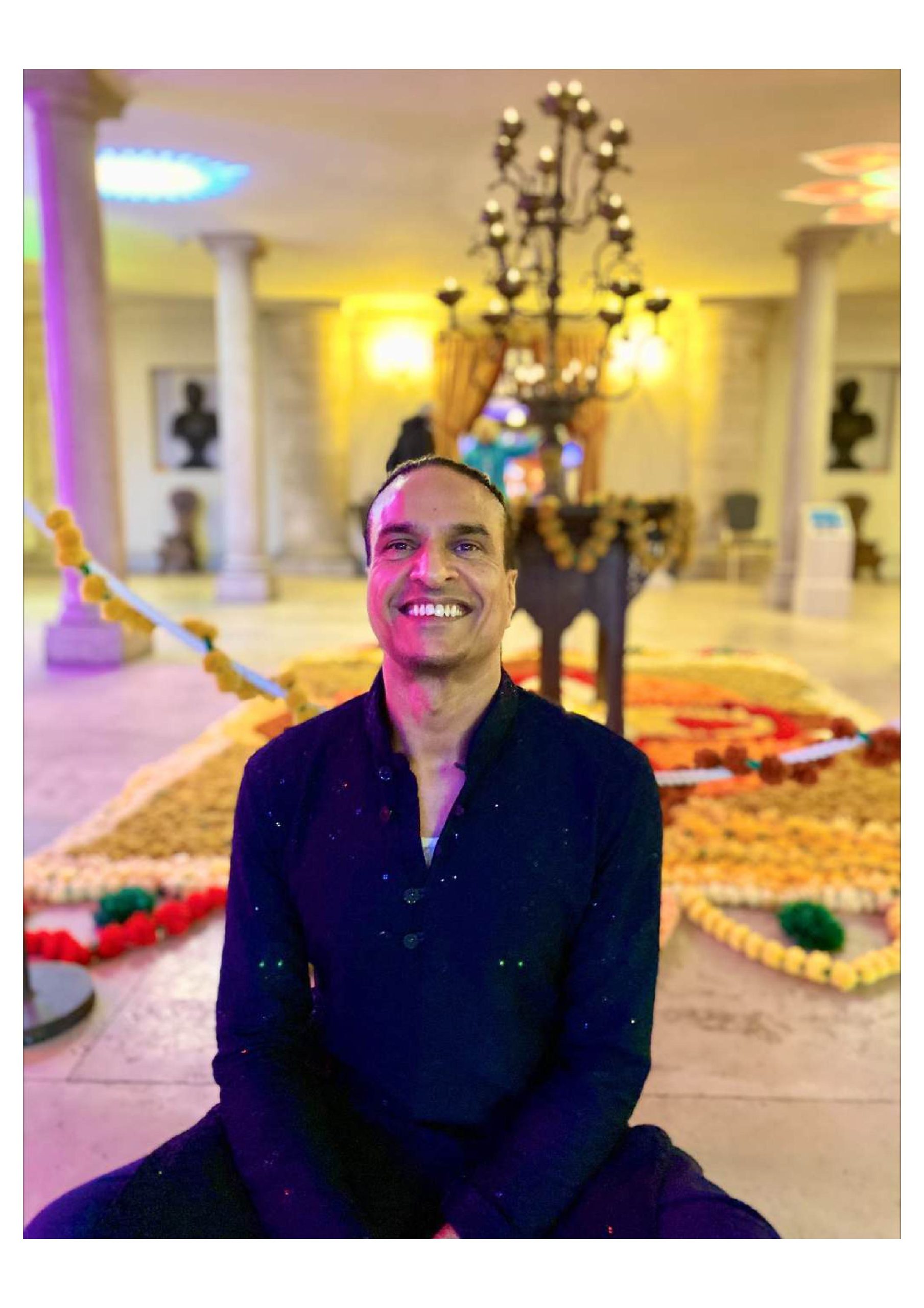 Mendi is sitting cross legged on the floor. His hair is tied back, he is smiling and is wearing a dark blue outfit. The background is blurred but looks like a grand stately home room with pillars and a candelabra. There are flower garlands decorating the floor behind Mendi
