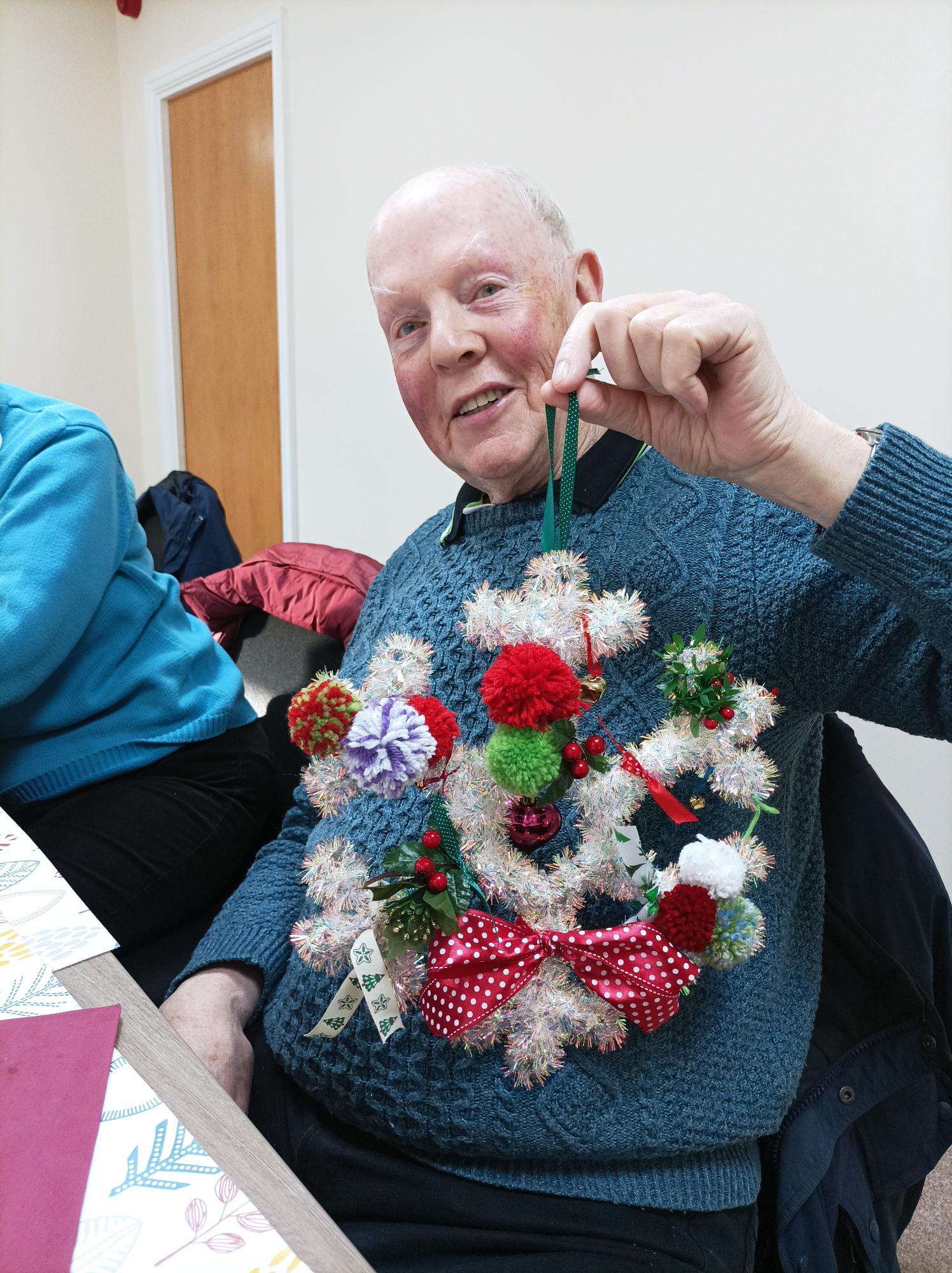 An older gentleman is proudly holding up a decorated snowflake shaped wreath covered in pom poms and ribbons