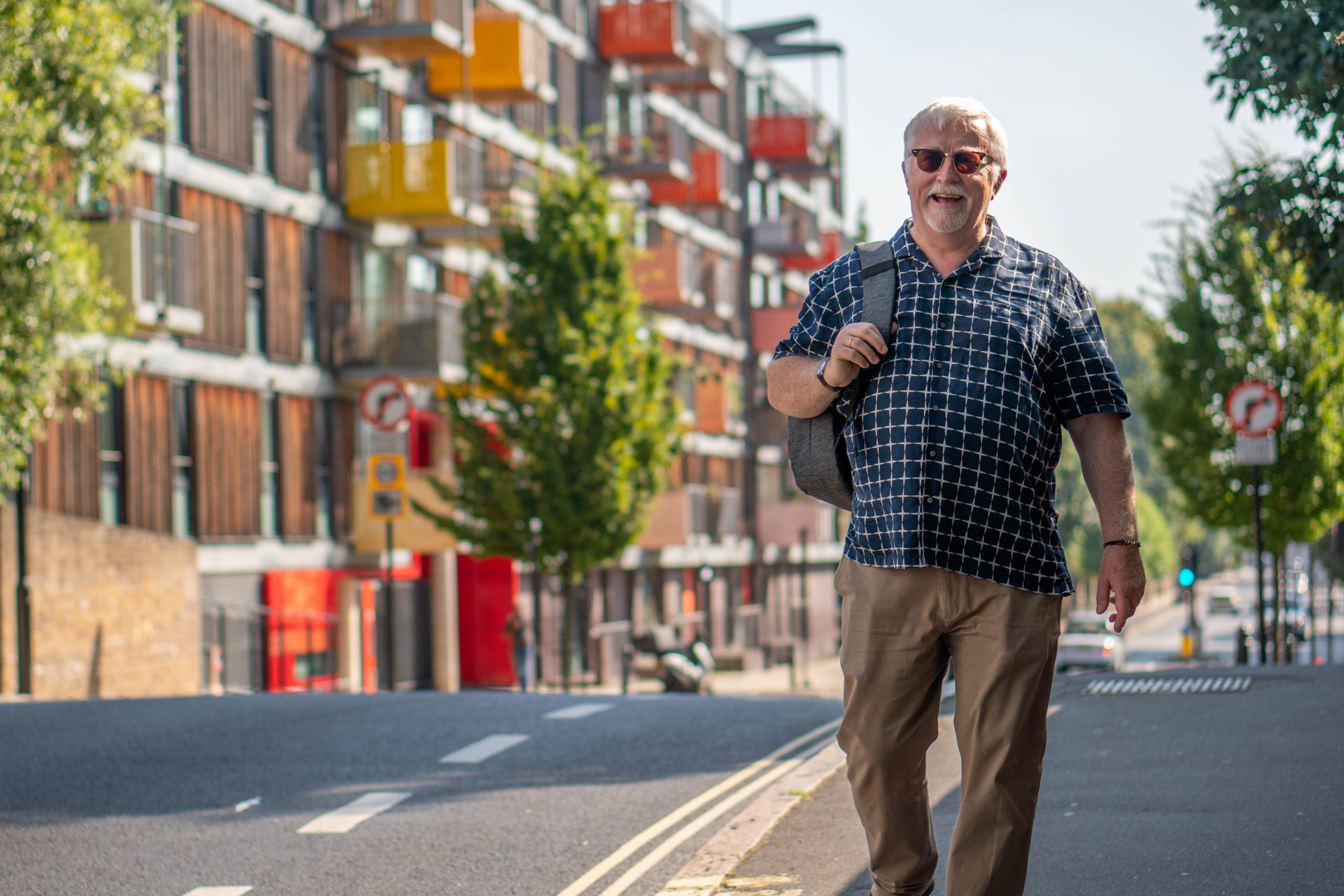 An older gentleman is walking down a city street with a backpack over his shoulder. The sun is shining, he had his sunglasses on and is smiling.