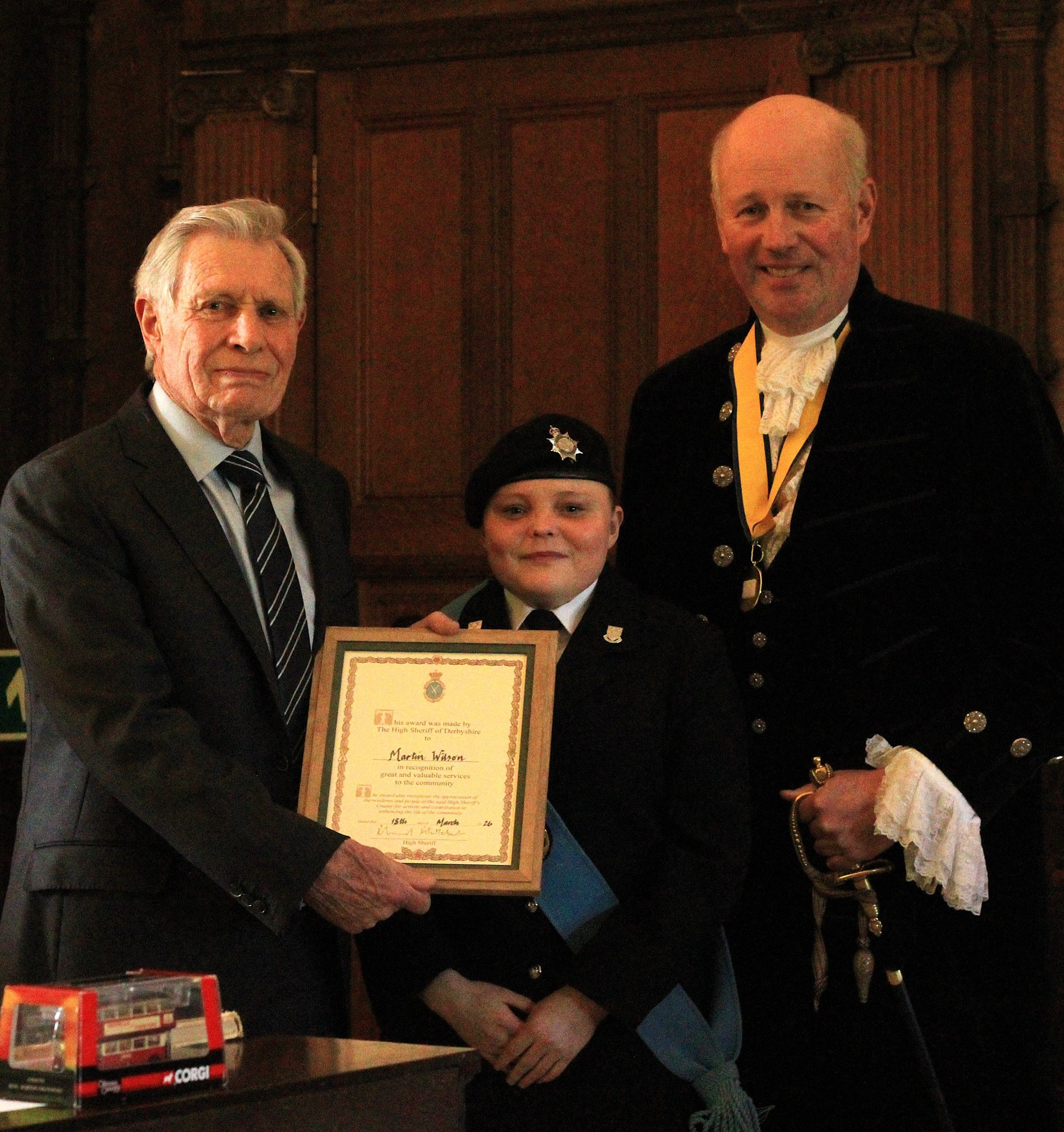 An older lady is sitting on a park bench with a laptop in front of her on a wooden table. There are trees in the background. She is wearing a blue blouse with small white spots, a gold watch and glasses. She is holding a mobile phone to her ear and her other hand is on the laptop. She has short grey hair and is smiling.