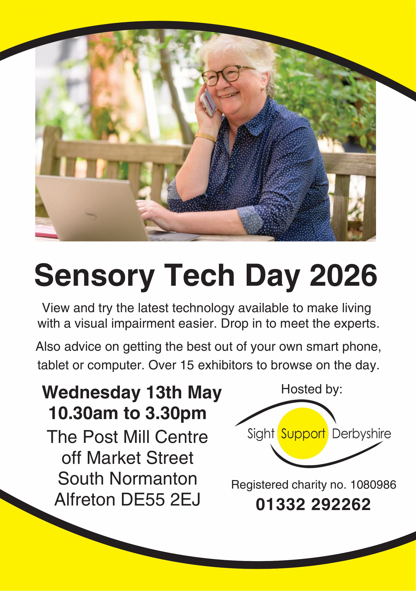 An older lady is sitting on a park bench with a laptop in front of her on a wooden table. There are trees in the background. She is wearing a blue blouse with small white spots, a gold watch and glasses. She is holding a mobile phone to her ear and her other hand is on the laptop. She has short grey hair and is smiling.