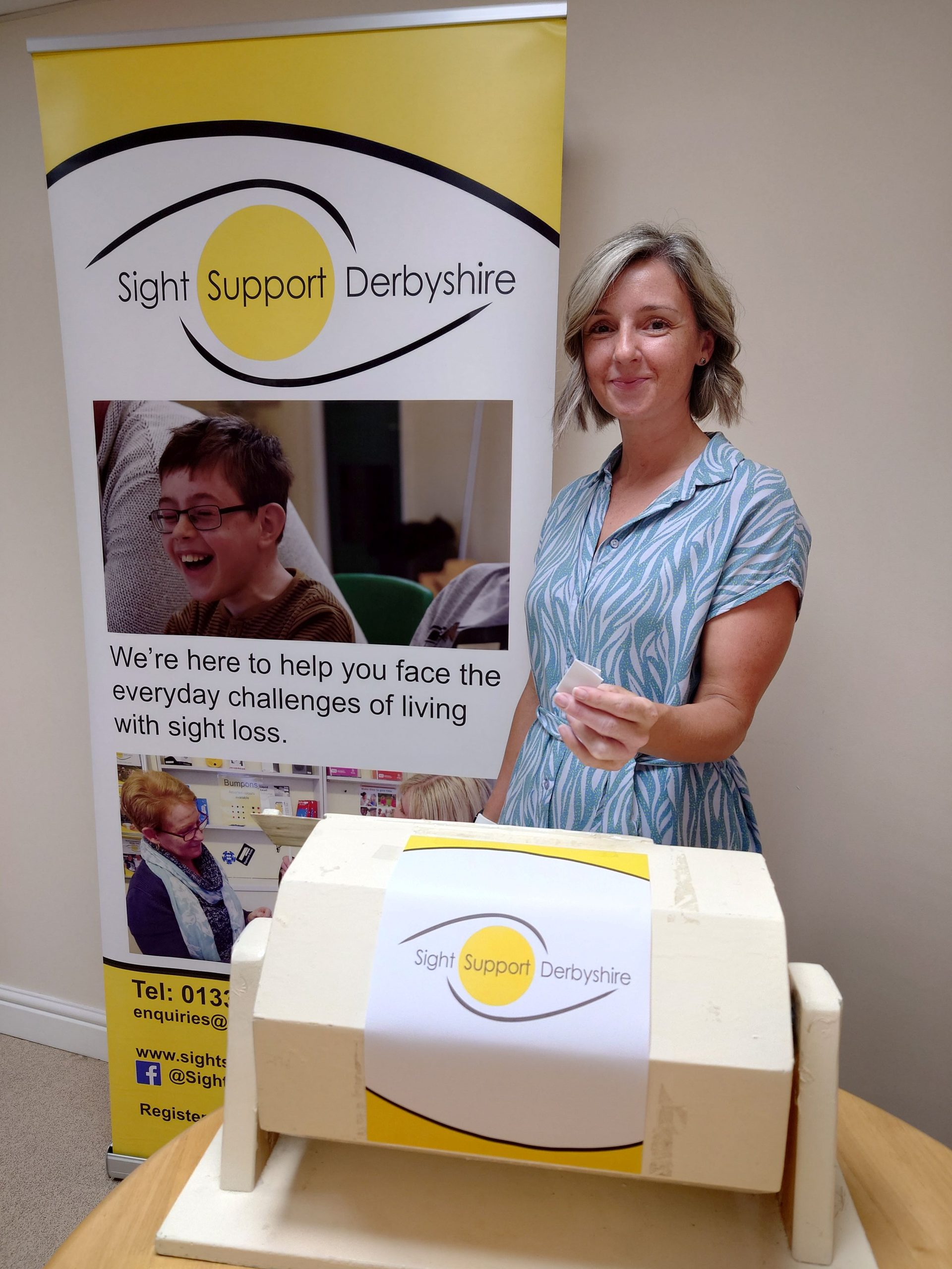 A lady is standing behind a tombola drum holding up a folded ticket. Behind her is a Sight Support Derbyshire banner